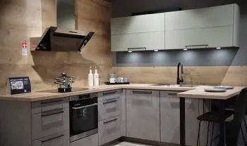 Moderne L-Küche mit grauen Fronten, hellen Oberschränken und Holzrückwand, ausgestattet mit Backofen, Kochfeld, Spüle, schwarzer Armatur sowie kleinem Sitzplatz am Tresen.